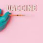 vaccine text and a person wearing latex glove while holding a syringe on pink background